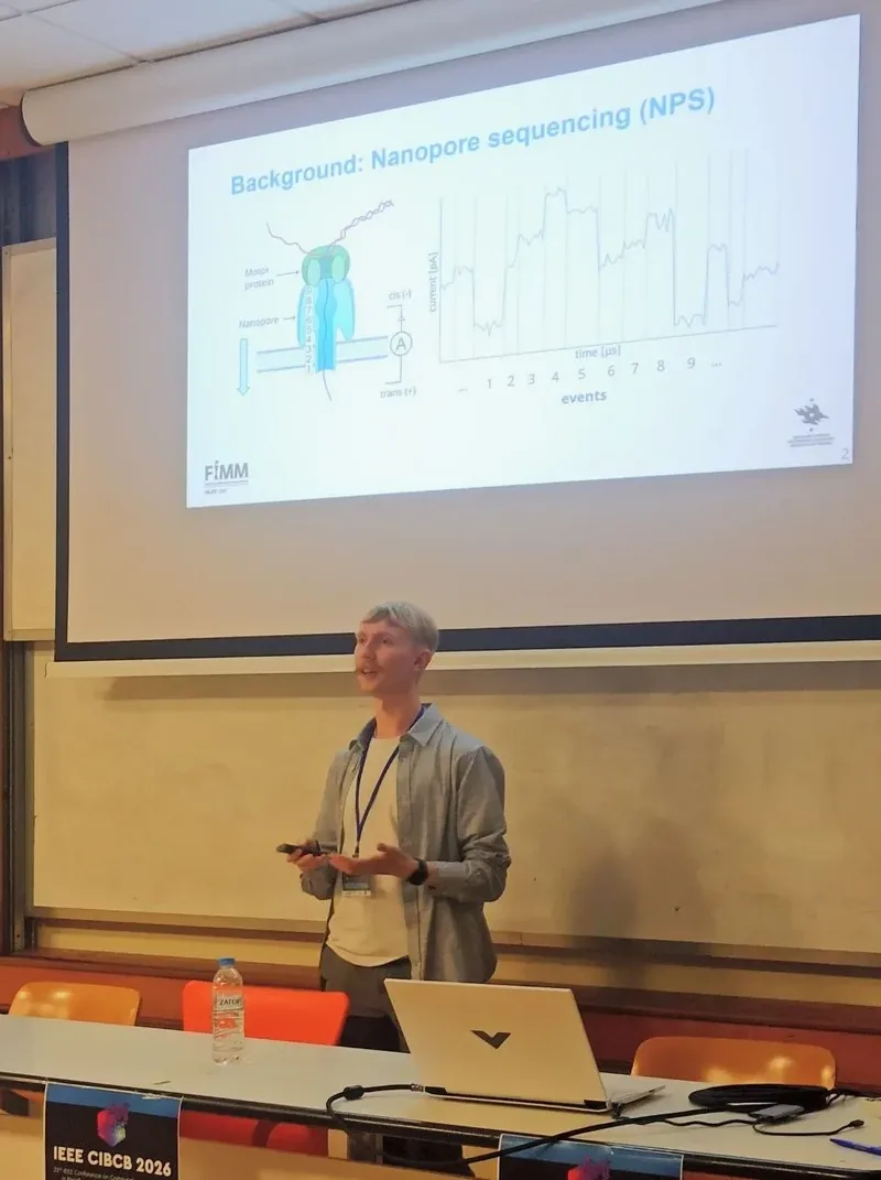 A speaker standing beside a projected slide headed "Background: Nanopore sequencing (NPS)", showing a diagram of a motor protein and a nanopore beside a current trace; a banner on the desk reads IEEE CIBCB 2026