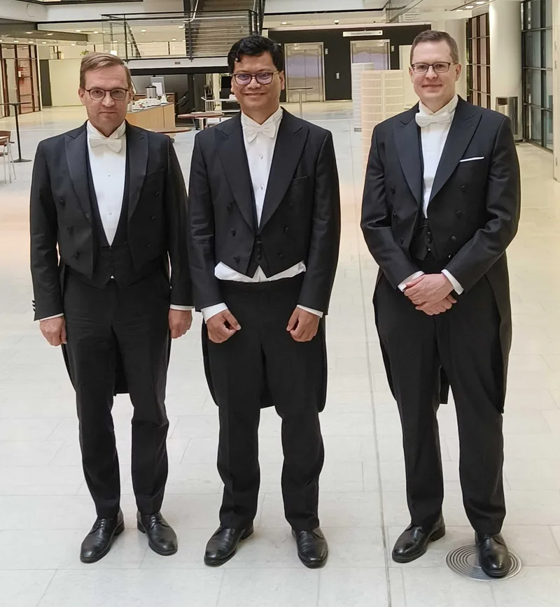 Three men in white-tie tailcoats standing side by side in the bright, tiled lobby of Biomedicum Helsinki
