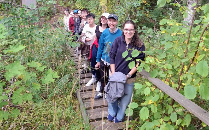 Ten members of the group standing in single file on a wooden boardwalk through dense green forest, looking up towards the camera